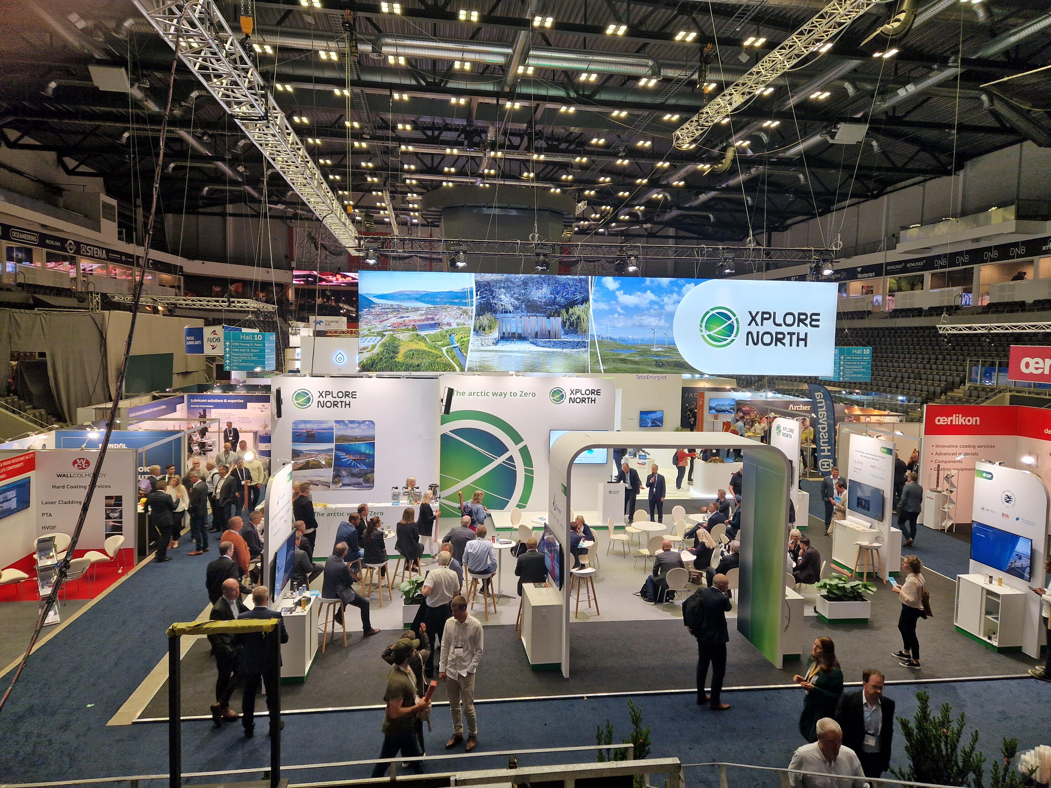 Standomr&aring;det til Xplore North under en stor messe, med mange bes&oslash;kende i samtaler rundt ulike utstillingspunkter. Store skjermer viser arktiske landskap, og standen er preget av gr&oslash;nn og hvit profilering.&rdquo;